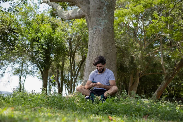 Güzel güneşli bir günde, cep telefonuyla parkta genç bir adam. Çalışmak ve rahatlamak için cep telefonu. Merkez ofis konsepti. Yeşil ve doğa geçmişi. Yüksek kalite fotoğraf