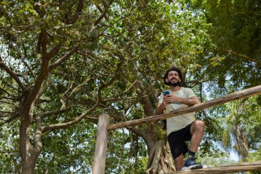 Güzel güneşli bir günde, cep telefonuyla parkta genç bir adam. Çalışmak ve rahatlamak için cep telefonu. Merkez ofis konsepti. Yeşil ve doğa geçmişi. Yüksek kalite fotoğraf