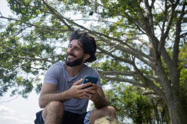 Güzel güneşli bir günde, cep telefonuyla parkta genç bir adam. Çalışmak ve rahatlamak için cep telefonu. Merkez ofis konsepti. Yeşil ve doğa geçmişi. Yüksek kalite fotoğraf