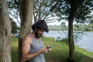 Güzel güneşli bir günde, cep telefonuyla parkta genç bir adam. Çalışmak ve rahatlamak için cep telefonu. Merkez ofis konsepti. Yeşil ve doğa geçmişi. Yüksek kalite fotoğraf