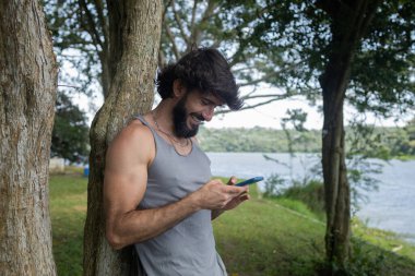 Güzel güneşli bir günde, cep telefonuyla parkta genç bir adam. Çalışmak ve rahatlamak için cep telefonu. Merkez ofis konsepti. Yeşil ve doğa geçmişi. Yüksek kalite fotoğraf