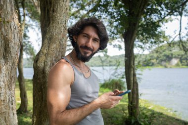 Güzel güneşli bir günde, cep telefonuyla parkta genç bir adam. Çalışmak ve rahatlamak için cep telefonu. Merkez ofis konsepti. Yeşil ve doğa geçmişi. Yüksek kalite fotoğraf
