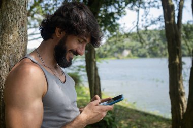 Güzel güneşli bir günde, cep telefonuyla parkta genç bir adam. Çalışmak ve rahatlamak için cep telefonu. Merkez ofis konsepti. Yeşil ve doğa geçmişi. Yüksek kalite fotoğraf