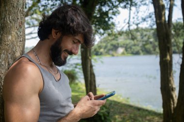 Güzel güneşli bir günde, cep telefonuyla parkta genç bir adam. Çalışmak ve rahatlamak için cep telefonu. Merkez ofis konsepti. Yeşil ve doğa geçmişi. Yüksek kalite fotoğraf