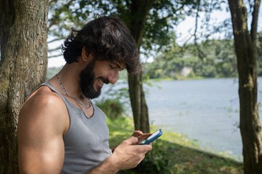 Güzel güneşli bir günde, cep telefonuyla parkta genç bir adam. Çalışmak ve rahatlamak için cep telefonu. Merkez ofis konsepti. Yeşil ve doğa geçmişi. Yüksek kalite fotoğraf