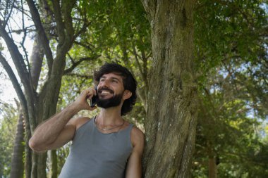 Güzel güneşli bir günde, cep telefonuyla parkta genç bir adam. Çalışmak ve rahatlamak için cep telefonu. Merkez ofis konsepti. Yeşil ve doğa geçmişi. Yüksek kalite fotoğraf