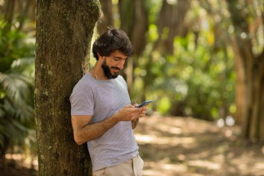 Gündüz vakti arka planda yeşil park olan akıllı telefon kullanan genç bir adamın görüntüsü. Cep telefonu, teknoloji, şehir konsepti. Yüksek kalite fotoğraf