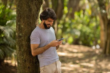 Gündüz vakti arka planda yeşil park olan akıllı telefon kullanan genç bir adamın görüntüsü. Cep telefonu, teknoloji, şehir konsepti. Yüksek kalite fotoğraf