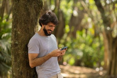 Gündüz vakti arka planda yeşil park olan akıllı telefon kullanan genç bir adamın görüntüsü. Cep telefonu, teknoloji, şehir konsepti. Yüksek kalite fotoğraf