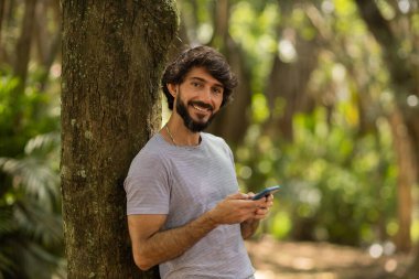 Gündüz vakti arka planda yeşil park olan akıllı telefon kullanan genç bir adamın görüntüsü. Cep telefonu, teknoloji, şehir konsepti. Yüksek kalite fotoğraf