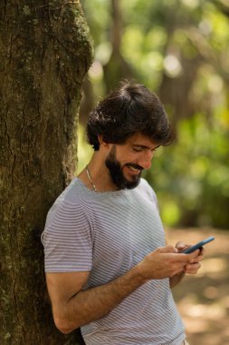 Gündüz vakti arka planda yeşil park olan akıllı telefon kullanan genç bir adamın görüntüsü. Cep telefonu, teknoloji, şehir konsepti. Yüksek kalite fotoğraf