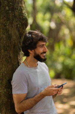 Gündüz vakti arka planda yeşil park olan akıllı telefon kullanan genç bir adamın görüntüsü. Cep telefonu, teknoloji, şehir konsepti. Yüksek kalite fotoğraf