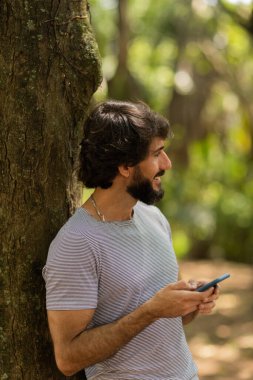 Gündüz vakti arka planda yeşil park olan akıllı telefon kullanan genç bir adamın görüntüsü. Cep telefonu, teknoloji, şehir konsepti. Yüksek kalite fotoğraf