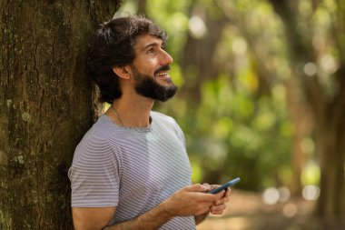 Gündüz vakti arka planda yeşil park olan akıllı telefon kullanan genç bir adamın görüntüsü. Cep telefonu, teknoloji, şehir konsepti. Yüksek kalite fotoğraf