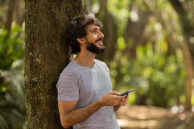 Gündüz vakti arka planda yeşil park olan akıllı telefon kullanan genç bir adamın görüntüsü. Cep telefonu, teknoloji, şehir konsepti. Yüksek kalite fotoğraf