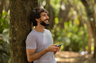 Gündüz vakti arka planda yeşil park olan akıllı telefon kullanan genç bir adamın görüntüsü. Cep telefonu, teknoloji, şehir konsepti. Yüksek kalite fotoğraf