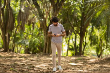 Gündüz vakti arka planda yeşil park olan akıllı telefon kullanan genç bir adamın görüntüsü. Cep telefonu, teknoloji, şehir konsepti. Yüksek kalite fotoğraf