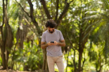Gündüz vakti arka planda yeşil park olan akıllı telefon kullanan genç bir adamın görüntüsü. Cep telefonu, teknoloji, şehir konsepti. Yüksek kalite fotoğraf