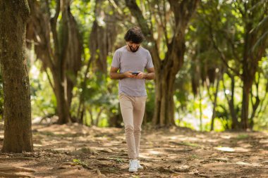 Gündüz vakti arka planda yeşil park olan akıllı telefon kullanan genç bir adamın görüntüsü. Cep telefonu, teknoloji, şehir konsepti. Yüksek kalite fotoğraf