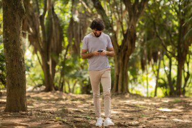 Gündüz vakti arka planda yeşil park olan akıllı telefon kullanan genç bir adamın görüntüsü. Cep telefonu, teknoloji, şehir konsepti. Yüksek kalite fotoğraf