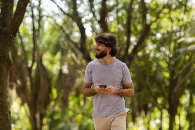 Gündüz vakti arka planda yeşil park olan akıllı telefon kullanan genç bir adamın görüntüsü. Cep telefonu, teknoloji, şehir konsepti. Yüksek kalite fotoğraf