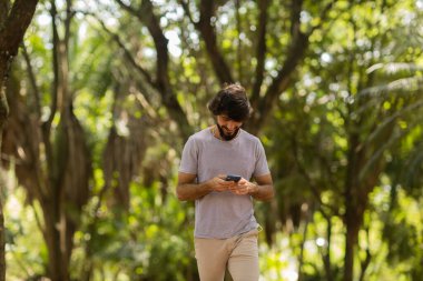 Gündüz vakti arka planda yeşil park olan akıllı telefon kullanan genç bir adamın görüntüsü. Cep telefonu, teknoloji, şehir konsepti. Yüksek kalite fotoğraf