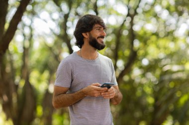 Gündüz vakti arka planda yeşil park olan akıllı telefon kullanan genç bir adamın görüntüsü. Cep telefonu, teknoloji, şehir konsepti. Yüksek kalite fotoğraf