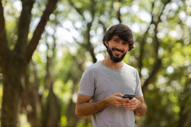 Gündüz vakti arka planda yeşil park olan akıllı telefon kullanan genç bir adamın görüntüsü. Cep telefonu, teknoloji, şehir konsepti. Yüksek kalite fotoğraf