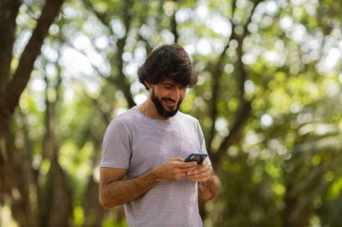 Gündüz vakti arka planda yeşil park olan akıllı telefon kullanan genç bir adamın görüntüsü. Cep telefonu, teknoloji, şehir konsepti. Yüksek kalite fotoğraf