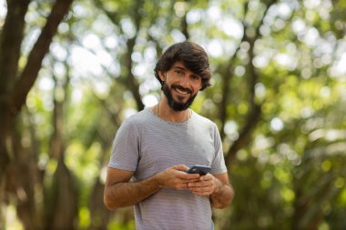 Gündüz vakti arka planda yeşil park olan akıllı telefon kullanan genç bir adamın görüntüsü. Cep telefonu, teknoloji, şehir konsepti. Yüksek kalite fotoğraf
