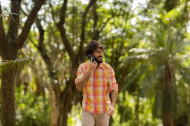 Gündüz vakti arka planda yeşil park olan akıllı telefon kullanan genç bir adamın görüntüsü. Cep telefonu, teknoloji, şehir konsepti. Yüksek kalite fotoğraf