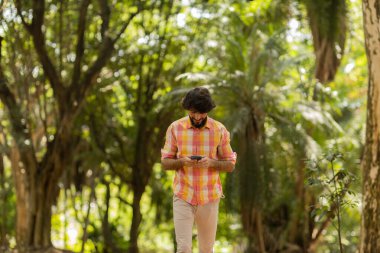 Gündüz vakti arka planda yeşil park olan akıllı telefon kullanan genç bir adamın görüntüsü. Cep telefonu, teknoloji, şehir konsepti. Yüksek kalite fotoğraf