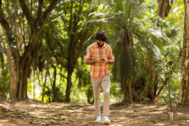 Gündüz vakti arka planda yeşil park olan akıllı telefon kullanan genç bir adamın görüntüsü. Cep telefonu, teknoloji, şehir konsepti. Yüksek kalite fotoğraf