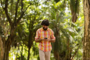 Gündüz vakti arka planda yeşil park olan akıllı telefon kullanan genç bir adamın görüntüsü. Cep telefonu, teknoloji, şehir konsepti. Yüksek kalite fotoğraf