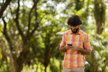 Gündüz vakti arka planda yeşil park olan akıllı telefon kullanan genç bir adamın görüntüsü. Cep telefonu, teknoloji, şehir konsepti. Yüksek kalite fotoğraf
