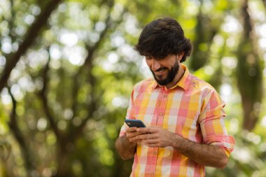 Gündüz vakti arka planda yeşil park olan akıllı telefon kullanan genç bir adamın görüntüsü. Cep telefonu, teknoloji, şehir konsepti. Yüksek kalite fotoğraf