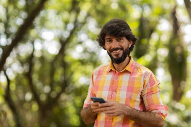 Gündüz vakti arka planda yeşil park olan akıllı telefon kullanan genç bir adamın görüntüsü. Cep telefonu, teknoloji, şehir konsepti. Yüksek kalite fotoğraf
