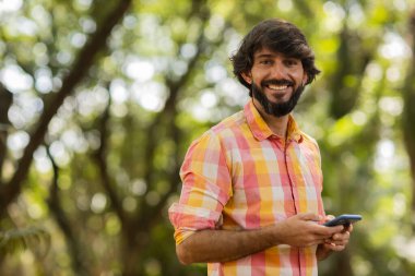 Gündüz vakti arka planda yeşil park olan akıllı telefon kullanan genç bir adamın görüntüsü. Cep telefonu, teknoloji, şehir konsepti. Yüksek kalite fotoğraf