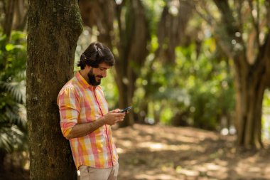 Gündüz vakti arka planda yeşil park olan akıllı telefon kullanan genç bir adamın görüntüsü. Cep telefonu, teknoloji, şehir konsepti. Yüksek kalite fotoğraf