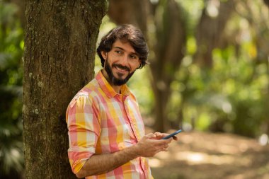 Gündüz vakti arka planda yeşil park olan akıllı telefon kullanan genç bir adamın görüntüsü. Cep telefonu, teknoloji, şehir konsepti. Yüksek kalite fotoğraf