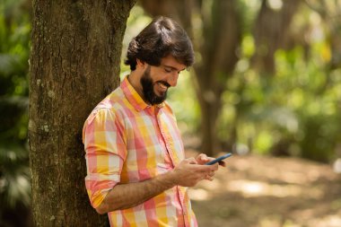Gündüz vakti arka planda yeşil park olan akıllı telefon kullanan genç bir adamın görüntüsü. Cep telefonu, teknoloji, şehir konsepti. Yüksek kalite fotoğraf