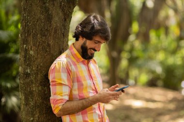 Gündüz vakti arka planda yeşil park olan akıllı telefon kullanan genç bir adamın görüntüsü. Cep telefonu, teknoloji, şehir konsepti. Yüksek kalite fotoğraf