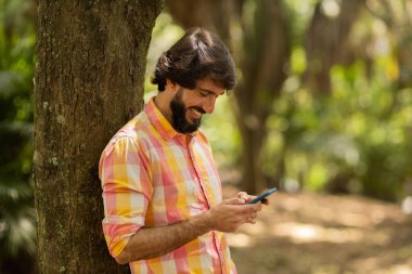 Gündüz vakti arka planda yeşil park olan akıllı telefon kullanan genç bir adamın görüntüsü. Cep telefonu, teknoloji, şehir konsepti. Yüksek kalite fotoğraf