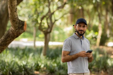 Gündüz vakti arka planda yeşil park olan akıllı telefon kullanan genç bir adamın görüntüsü. Cep telefonu, teknoloji, şehir konsepti. Yüksek kalite fotoğraf