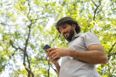 Gündüz vakti arka planda yeşil park olan akıllı telefon kullanan genç bir adamın görüntüsü. Cep telefonu, teknoloji, şehir konsepti. Yüksek kalite fotoğraf
