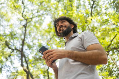Gündüz vakti arka planda yeşil park olan akıllı telefon kullanan genç bir adamın görüntüsü. Cep telefonu, teknoloji, şehir konsepti. Yüksek kalite fotoğraf