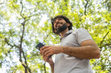 Gündüz vakti arka planda yeşil park olan akıllı telefon kullanan genç bir adamın görüntüsü. Cep telefonu, teknoloji, şehir konsepti. Yüksek kalite fotoğraf