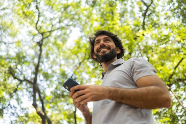 Gündüz vakti arka planda yeşil park olan akıllı telefon kullanan genç bir adamın görüntüsü. Cep telefonu, teknoloji, şehir konsepti. Yüksek kalite fotoğraf