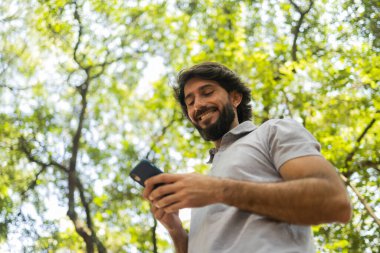Gündüz vakti arka planda yeşil park olan akıllı telefon kullanan genç bir adamın görüntüsü. Cep telefonu, teknoloji, şehir konsepti. Yüksek kalite fotoğraf