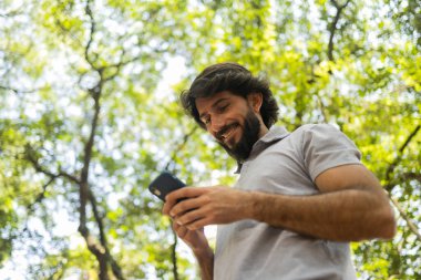 Gündüz vakti arka planda yeşil park olan akıllı telefon kullanan genç bir adamın görüntüsü. Cep telefonu, teknoloji, şehir konsepti. Yüksek kalite fotoğraf